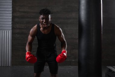 Güçlü ve kendinden emin bir Afro-Amerikan boksör kırmızı eldivenler giyiyor, atletik yapısını ve kararlılığını gösteriyor, bağırıyor ve spor salonunun karanlık, minimalist atmosferinde saldırganlığını ifade ediyor..