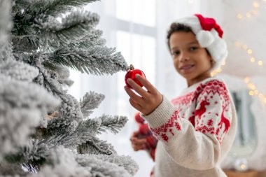 Afrika kökenli Amerikalı Noel Baba şapkalı ve kışlık süveterli genç çocuk Noel ağacını evde kırmızı topla süslüyor, çocuk yeni yılı ve Noel 'i kutluyor ve oyuncak süsleme yapıyor.