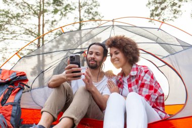 Genç Afro-Amerikan çift kamp ekipmanları ve sırt çantalarıyla ormanda çadırda oturuyorlar ve akıllı telefon kullanıyorlar, kadın ve erkek kamp yapıyorlar ve internetten cep telefonlarını izliyorlar.