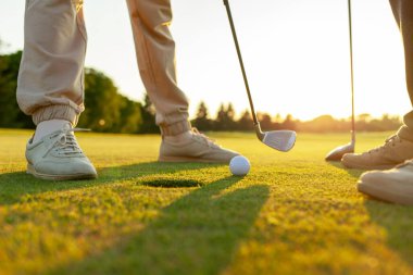 Gün batımında golf oynayan erkek ve kadın, golf topuna ve sopalara yakın çekim, yeşil çimlerin üzerinde insanların ayakları, oyunda yarışıyor ve deliğe vuruyor.