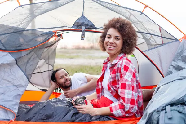 Genç Afro-Amerikan çift ormanda bir çadırda çay içerek yürüyüş yapıyorlar ve gülümsüyorlar, kadın ve erkek çadırlarla seyahat ediyorlar ve kahve içiyorlar ve aktif olarak şehir dışında dinleniyorlar.