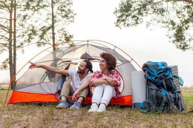 Genç Afro-Amerikan çift kamp ekipmanları ve sırt çantalarıyla ormanda çadırda oturuyorlar ve elleriyle işaret ediyorlar, erkek ve kadın kamp alanındaki çadırlarla dinleniyorlar ve