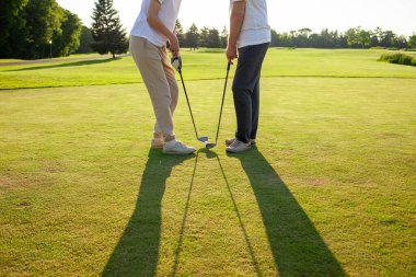 Gün batımında golf oynayan erkek ve kadın, golf topuna ve sopalara yakın çekim, yeşil çimlerin üzerinde insanların ayakları, oyunda yarışıyor ve deliğe vuruyor.