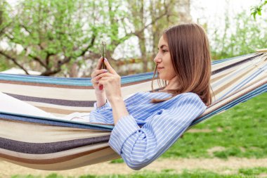 Genç neşeli kız yazın bahçede rahat bir hamakta uzanır ve internetten akıllı telefon kullanır. Parkta dinlenir ve cep telefonuna bakar ve gülümser.