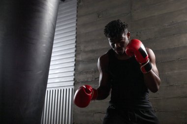 Kırmızı eldiven giyen güçlü Afrikalı Amerikalı boksör spor salonunda kum torbasına vurmaya hazırlanıyor. Boks müsabakasından önce atlet trenleri ve kararlılığını gösteriyor.