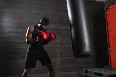 Genç, güçlü, kırmızı eldivenli Afrikalı Amerikalı boksör kum torbasına vuruyor, kaslı atlet trenleri koyu renk spor salonunda yumrukluyor ve siyah arka planda spor yapıyor.
