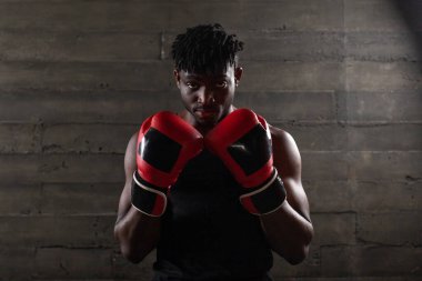 Kırmızı eldivenli güçlü Afro-Amerikan boksör bir kum torbasının yanında savunma pozisyonunda duruyor. Kırmızı eldivenli sporcu siyah arka plandaki kameraya bakıyor.