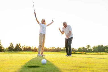 Yaşlı adam ve kadın golfün galibiyetini kutluyor ve el kaldırarak bağırıyor, yaşlı çift gün batımında golf oynuyor ve golf ekipmanlarıyla başarıyı kutluyor.