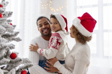 Noel Baba şapkalı Afro-Amerikan ailesi evde birlikte yeni yılı kutluyor ve Noel ağacını süslüyor, genç çocuk anne ve babasıyla yeni yılı kutluyor ve kırmızı oyuncak tutuyor.