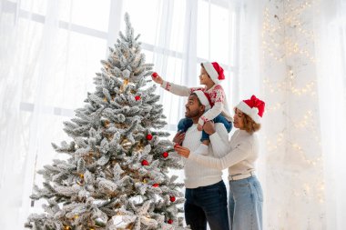 Noel Baba şapkalı Afro-Amerikan ailesi evde birlikte yeni yılı kutluyor ve Noel ağacını süslüyor, genç çocuk anne ve babasıyla yeni yılı kutluyor ve kırmızı oyuncak tutuyor.