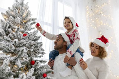 Noel Baba şapkalı Afro-Amerikan ailesi evde birlikte yeni yılı kutluyor ve Noel ağacını süslüyor, genç çocuk anne ve babasıyla yeni yılı kutluyor ve kırmızı oyuncak tutuyor.