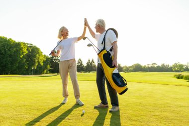 Üniformalı yaşlı çift golf oyununda zaferi ve başarıyı kutluyor ve gün batımında golf oynayan yaşlı adam ve kadına beşlik çakıyor ve açık hava sporları yapıyor.