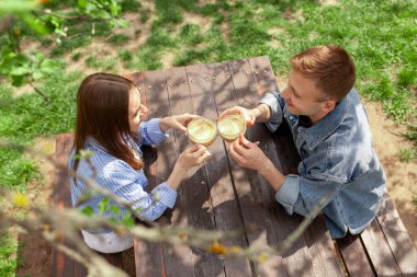 Kot elbiseli genç bir çift, tahta bir masada bahçede oturuyor ve yazın bir randevuda kahve içiyor, kız ve erkek açık hava kafede kahvaltı yapıyor ve sohbet ediyorlar.