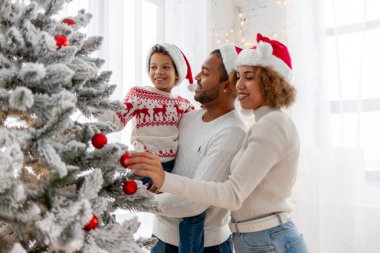 Noel Baba şapkalı Afro-Amerikan ailesi evde birlikte yeni yılı kutluyor ve Noel ağacını süslüyor, genç çocuk anne ve babasıyla yeni yılı kutluyor ve kırmızı oyuncak tutuyor.