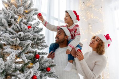 Noel Baba şapkalı Afro-Amerikan ailesi evde birlikte yeni yılı kutluyor ve Noel ağacını süslüyor, genç çocuk anne ve babasıyla yeni yılı kutluyor ve kırmızı oyuncak tutuyor.