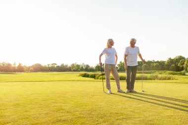 Güneş batarken golf sahasında kucaklaşan, beyaz üniformalı yaşlı ve yaşlı çift golf oynuyor ve doğada kameraya bakıp gülümsüyor.