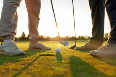 Gün batımında golf oynayan erkek ve kadın, golf topuna ve sopalara yakın çekim, yeşil çimlerin üzerinde insanların ayakları, oyunda yarışıyor ve deliğe vuruyor.