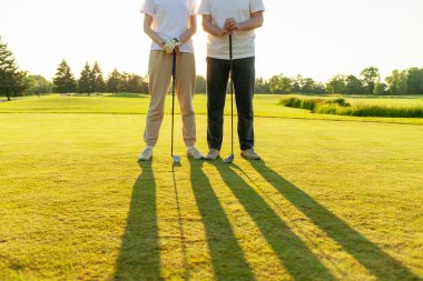 Gün batımında golf oynayan erkek ve kadın, golf topuna ve sopalara yakın çekim, yeşil çimlerin üzerinde insanların ayakları, oyunda yarışıyor ve deliğe vuruyor.
