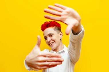 Genç, kısa saçlı, kızıl saçlı kız planları yapıyor ve sarı izole edilmiş bir arkaplanda elleri olan bir çerçeve gösteriyor. Piercing çerçeveli hipster bir kadın ve fotoğraf çekiyor.