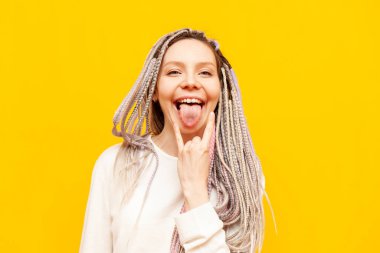 Renkli rastalı genç kız kaya jestleri ve sarı izole arka planda çığlıklar gösteriyor. Eşsiz saç stili ve örgüleri olan çılgın hippi kadın dil gösteriyor.