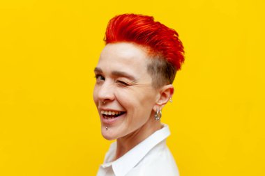 Genç, kısa saçlı, kızıl saçlı bir kız sarı izole bir arka planda gülümseyip göz kırparak flört ediyor.