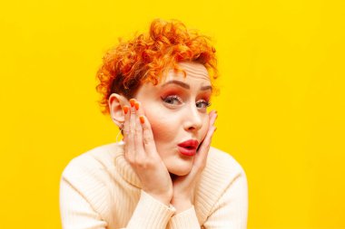 Genç, kızıl saçlı, kıvırcık saçlı, heyecanlı kadın şaşırır ve sarı izole bir arkaplanda kameraya bakar.