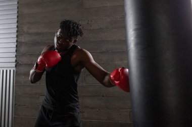 Genç, güçlü, kırmızı eldivenli Afrikalı Amerikalı boksör kum torbasına vuruyor, kaslı atlet trenleri koyu renk spor salonunda yumrukluyor ve siyah arka planda spor yapıyor.