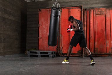 Genç, güçlü, kırmızı eldivenli Afrikalı Amerikalı boksör kum torbasına vuruyor, kaslı atlet trenleri koyu renk spor salonunda yumrukluyor ve siyah arka planda spor yapıyor.
