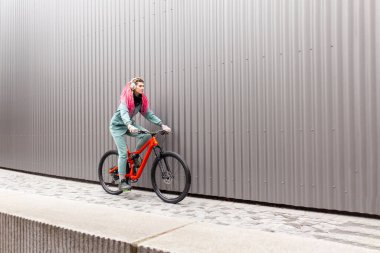 Kalın elbiseli genç hippi kız gri bir duvarın arkasında bisiklet sürüyor ve gülümsüyor, sonbaharda soğuk havada bisiklet süren öğrenci bisikletçi.