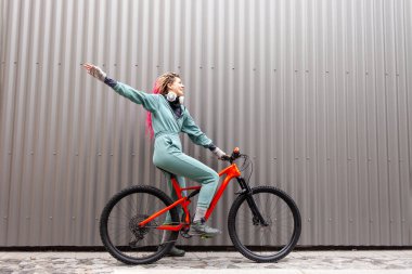 Kalın elbiseli genç hippi kız gri bir duvarın arkasında bisiklet sürüyor ve gülümsüyor, sonbaharda soğuk havada bisiklet süren öğrenci bisikletçi.