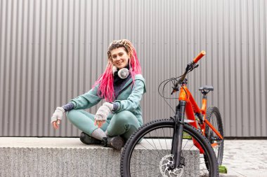 Kalın elbiseli genç hippi kız gri bir duvarın arkasında bisikletiyle oturuyor ve gülümsüyor. Öğrenci bisikletçi dinleniyor ve sonbaharda soğuk havada bekliyor.