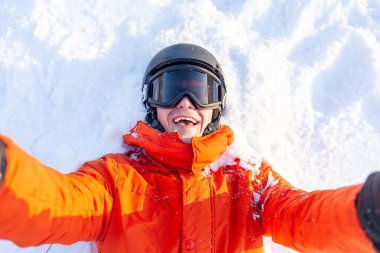 Kayak elbiseli genç adam kışın karda yatar ve selfie çeker, turuncu ceketli adam, miğfer ve gözlüklü adam tatil köyünde sevinir ve fotoğraf çeker.