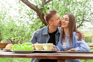 Kot elbiseli genç bir çift, ahşap masada bahçede oturuyor ve kahve içiyor ve yazın randevularında öpüşüyor, kız ve erkek açık hava kafede kahvaltı ediyor ve flört ediyor.