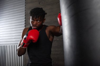 Genç, güçlü, kırmızı eldivenli Afrikalı Amerikalı boksör kum torbasına vuruyor, kaslı atlet trenleri koyu renk spor salonunda yumrukluyor ve siyah arka planda spor yapıyor.
