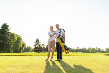 Üniformalı yaşlı çift ellerinde golf sopalarıyla gün batımında golf sahasının arka planında durup gülümseyen yaşlı adam ve kadın, golf oynarken sarılıp gülümsüyor ve fotokopiye bakıyorlardı.