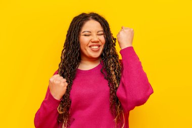 Rasta saçlı tombul Afrikalı Amerikalı kadın zaferi kutluyor ve sarı yalıtılmış arka planda başarı elde ediyor, artı at kuyruklu cüssesi olan kız iyi şansa seviniyor.