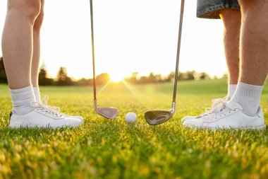 Üniformalı genç bir çift gün batımında çimenlikte golf oynuyor, eğitmenli bir kadın golf antrenmanı yapıyor ve aktif olarak dinleniyor.