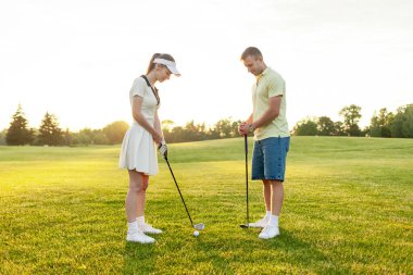 Üniformalı genç bir çift gün batımında çimenlikte golf oynuyor, eğitmenli bir kadın golf antrenmanı yapıyor ve aktif olarak dinleniyor.