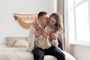 Genç ve mutlu bir çift evdeki yatakta sarılıyor ve gülümsüyor. Kadın erkeği sıcak yumuşak bir battaniyeyle örtüyor.