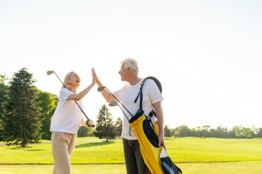 Üniformalı yaşlı çift golf oyununda zaferi ve başarıyı kutluyor ve gün batımında golf oynayan yaşlı adam ve kadına beşlik çakıyor ve açık hava sporları yapıyor.