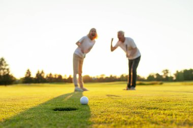 Sahada golf oynayan ve gün batımında zaferi kutlayan yaşlı bir çift yaşlı adam ve kadın golf topunu bir deliğe atıyorlar ve dışarıda birlikte başarıyla eğleniyorlar.