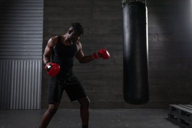 Genç, güçlü, kırmızı eldivenli Afrikalı Amerikalı boksör kum torbasına vuruyor, kaslı atlet trenleri koyu renk spor salonunda yumrukluyor ve siyah arka planda spor yapıyor.