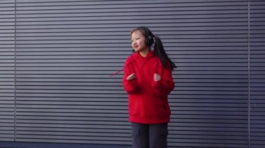 Kırmızı kapüşonlu genç kız kulaklıkla müzik dinliyor ve sokakta dans ediyor, enerjik kadın hip hop dansçısı şehrin arka planında duvarın arkasında dans ediyor.
