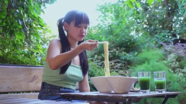 Genç, neşeli Asyalı bir kadın açık kafede çubuklarla ramen yiyor. Parktaki bir restoranda erişte yiyor.