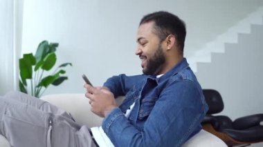 Neşeli Afrikalı Amerikalı adam akıllı telefon kullanıyor ve evde gülümsüyor, yetişkin adam sandalyeye uzanıyor ve mesaj yazıyor ve gülüyor.
