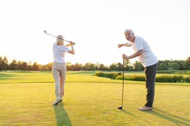 Yaşlı çift golf sahasında golf oynuyor ve gün batımında sohbet ediyor. Yaşlı erkek ve kadın dışarıda dinleniyor ve gülüyor. Emekliler tatilde.