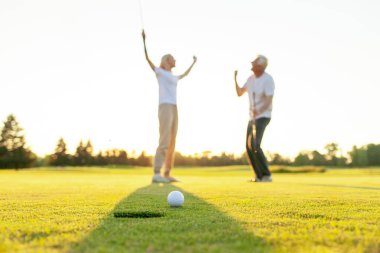 Sahada golf oynayan ve gün batımında zaferi kutlayan yaşlı bir çift yaşlı adam ve kadın golf topunu bir deliğe atıyorlar ve dışarıda birlikte başarıyla eğleniyorlar.