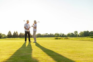Güneş batarken golf sahasında kucaklaşan, beyaz üniformalı yaşlı ve yaşlı çift golf oynuyor ve doğada kameraya bakıp gülümsüyor.
