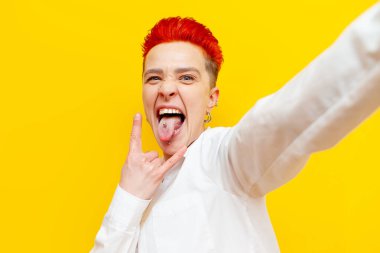 Genç, kısa saçlı, kızıl saçlı punk kız rock jesti sergiliyor ve sarı izole arka planda selfie çekiyor. Piercingli çılgın hippi kadın piercingli ve dilinde piercing olduğunu gösteriyor.