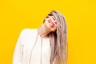 Siyah saçlı, neşeli genç bir kız sarı izole bir arka planda gülümsüyor ve seviniyor. Eşsiz saç stili ve örgüleriyle gözlerini kapatan hippi bir kadın.